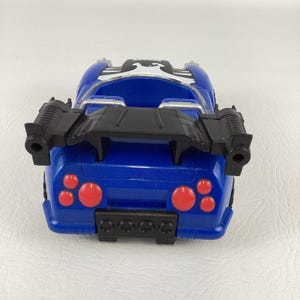 May include: A blue toy car with a black spoiler and red taillights. The car has a white and black design on the top and two black side attachments. The toy car is on a white surface.