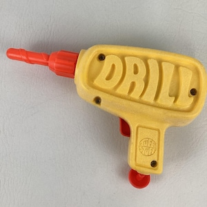 Pode incluir: Uma furadeira de brinquedo de plástico amarelo com uma broca de plástico vermelha. A palavra DRILL está gravada em letras grandes na lateral do corpo amarelo. Um pequeno gatilho vermelho é visível e uma peça circular vermelha na base.