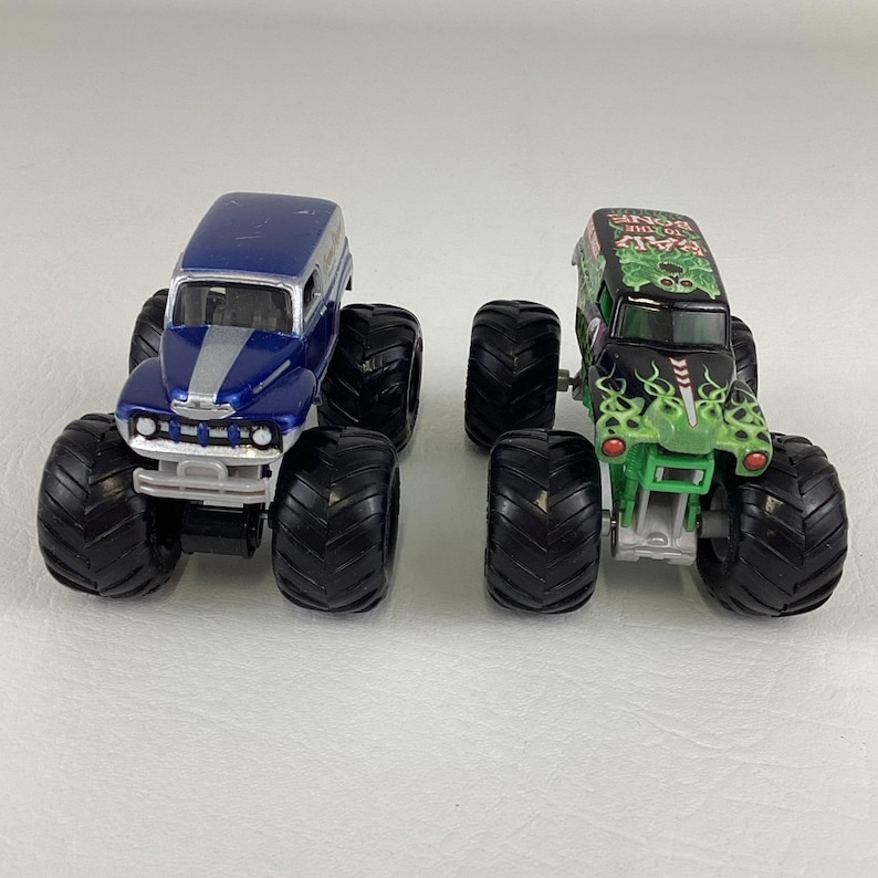May include: Two toy monster trucks. One is blue with silver stripes, the other is black with green flames and the text "Grave Digger". Both have large black tires and are on a white surface.