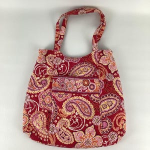 Puede incluir: Un bolso tote acolchado con un estampado de cachemira en tonos rojos, rosas y naranjas. El bolso tiene dos asas y un bolsillo delantero. El diseño de cachemira se repite en el exterior del bolso.