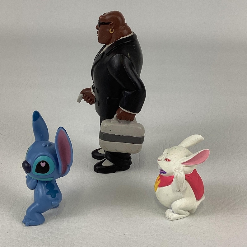 May include: A collection of cartoon character figurines. A blue alien-like creature with large ears and a heart-shaped eye, a white rabbit with pink ears and a red vest, and a man in a black suit holding a briefcase.