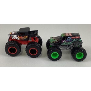May include: Two toy monster trucks. One is black and red with "Hot Wheels" text. The other is green and black with "Grave Digger" text and a skull graphic. Both have large black tires.