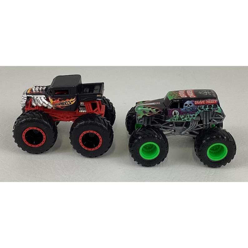 May include: Two toy monster trucks. One is black and red with "Hot Wheels" text. The other is green and black with "Grave Digger" text and a skull graphic. Both have large black tires.