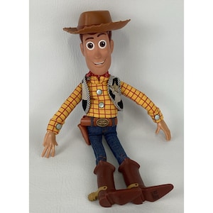 May include: A Woody action figure, featuring a brown cowboy hat, a yellow plaid shirt, a black and white vest, blue jeans, and brown cowboy boots. The figure has a red neckerchief and a brown belt with a gold buckle. The toy is a collectible item.