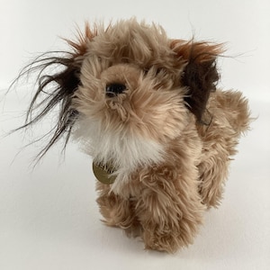 May include: A brown and white plush toy dog with a gold tag around its neck. The dog has long, shaggy fur and brown ears.