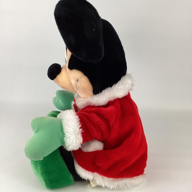 Puede incluir: Un peluche de Mickey Mouse que lleva una chaqueta roja con ribete de piel sint&eacute;tica blanca en el cuello y los pu&ntilde;os. El personaje tiene grandes orejas negras, cara color canela, nariz negra, y lleva manoplas verdes y zapatos verdes, visto de perfil.
