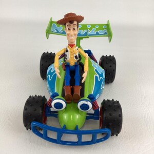 May include: A toy car featuring the Woody character from Toy Story. The car is primarily green with blue and white accents, black tires, and a blue front bumper. Woody is wearing a brown cowboy hat, yellow shirt, blue pants, and brown boots.