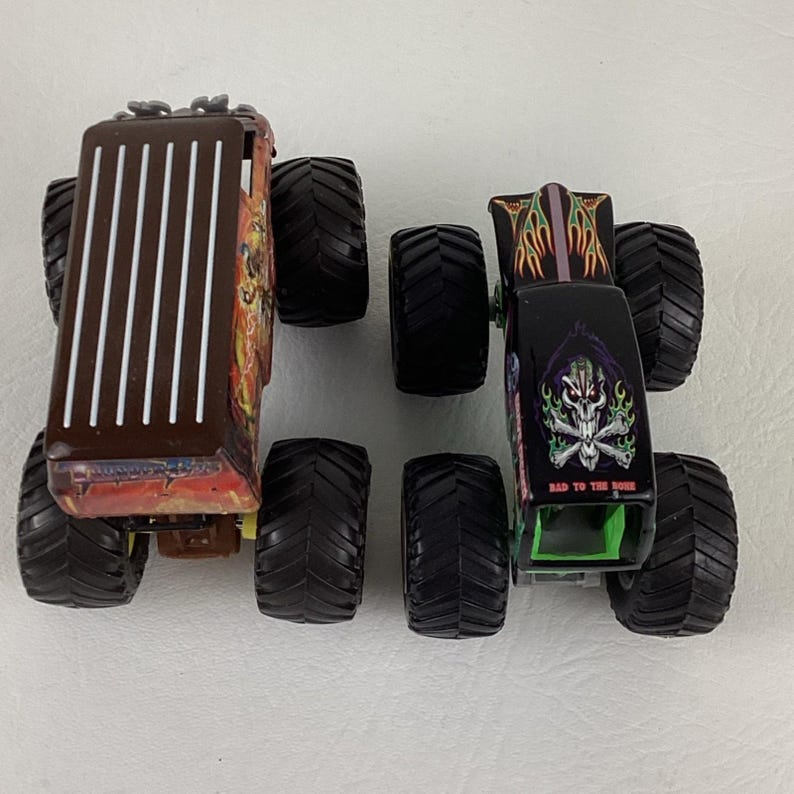 May include: Two toy monster trucks. One is brown with white vertical stripes and a detailed side graphic. The other is black with a skull graphic and the text "BAD TO THE BONE". Both have large black tires.