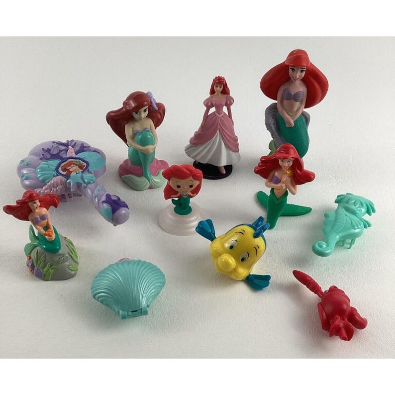 May include: A collection of Ariel-themed figurines and accessories. The set includes Ariel figures with red hair and mermaid tails, a Flounder figure, a seahorse, a shell-shaped compact, and other character-themed items.