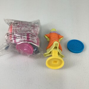 May include: A collection of McDonald's Happy Meal toys. One toy is a figure in an orange robe with a yellow base and a blue disc. Another toy is still in its original packaging. The packaging has text in multiple languages.