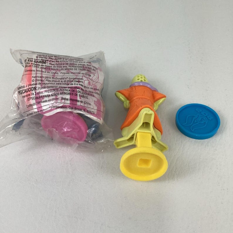 May include: A collection of McDonald's Happy Meal toys. One toy is a figure in an orange robe with a yellow base and a blue disc. Another toy is still in its original packaging. The packaging has text in multiple languages.