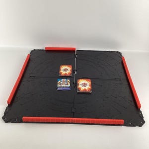Peut inclure: Un tapis de jeu pliable noir et rouge avec un motif circulaire. Le tapis est en plastique et a une surface texturée. Il y a trois cartes sur le tapis, une avec le texte "RULEBOOK" et deux avec le texte "BAKUGAN".
