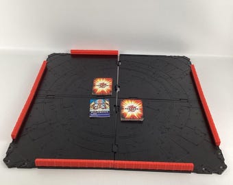 Pode incluir: Um tapete de jogo dobrável preto e vermelho com um design circular. O tapete é feito de plástico e tem uma superfície texturizada. Há três cartas no tapete, uma com o texto "RULEBOOK" e duas com o texto "BAKUGAN".