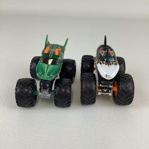 Puede incluir: Dos camiones monstruo de juguete. Uno es verde con un dise&ntilde;o de drag&oacute;n, el otro es blanco con un dise&ntilde;o de tibur&oacute;n. Ambos tienen neum&aacute;ticos negros grandes y detalles en naranja. Los camiones est&aacute;n dise&ntilde;ados para ni&ntilde;os.
