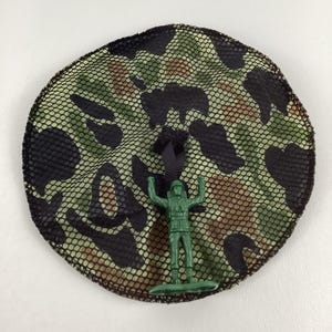 May include: A round camouflage-patterned net with a black border and a black ribbon. A green plastic toy soldier stands in front of the net, arms raised. The net features a pattern of green, brown, and black shapes.