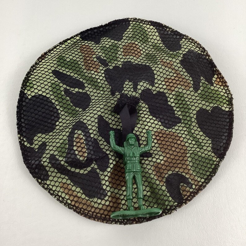 May include: A round camouflage-patterned net with a black border and a black ribbon. A green plastic toy soldier stands in front of the net, arms raised. The net features a pattern of green, brown, and black shapes.