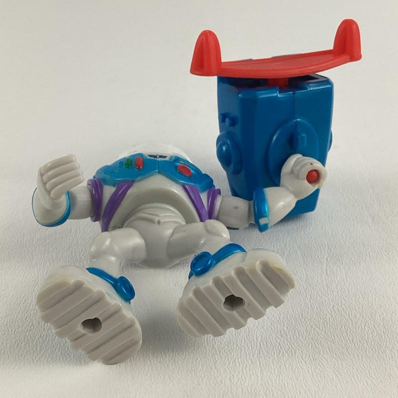Op de afbeelding: Een gedemonteerde Buzz Lightyear speelgoedfiguur. De figuur is voornamelijk wit met blauwe, paarse en rode accenten. De losgemaakte blauwe rugzak heeft een rode vleugelachtige structuur bovenop. Het speelgoed ligt op een witte ondergrond.