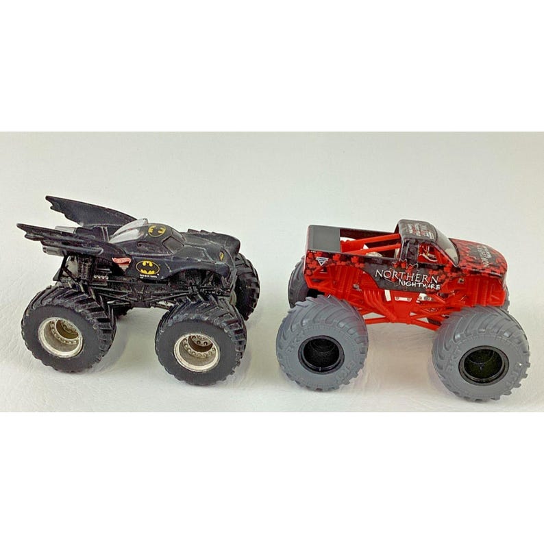 May include: Two toy monster trucks. One is black with a Batman logo and wing-like features. The other is red with a black and red pattern and the words "Northern Nightmare". Both have large, gray tires.