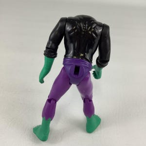 Puede incluir: Un torso de figura de acci&oacute;n sin cabeza con parte superior del cuerpo negra, secci&oacute;n de cintura y cadera morada con una ranura rectangular, y antebrazos y parte inferior de las piernas verdes. La figura tiene articulaciones en hombros, caderas y rodillas, mostrando una estructura muscular moldeada.