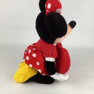 Pu&ograve; includere: Una bambola di peluche di Minnie Mouse seduta. Minnie indossa un vestito rosso a pois bianchi, un fiocco abbinato e scarpe nere. La bambola ha orecchie nere e una borsetta rossa a forma di cuore.