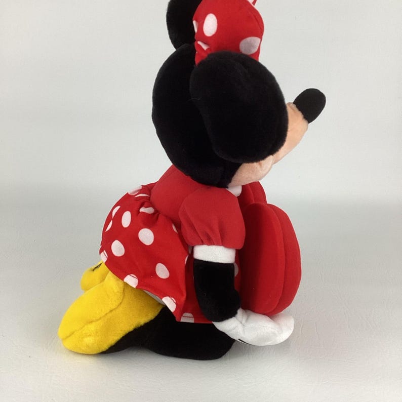 Pu&ograve; includere: Una bambola di peluche di Minnie Mouse seduta. Minnie indossa un vestito rosso a pois bianchi, un fiocco abbinato e scarpe nere. La bambola ha orecchie nere e una borsetta rossa a forma di cuore.
