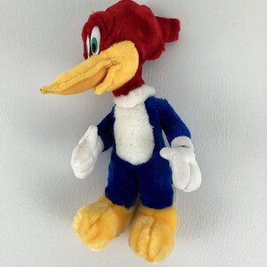Op de afbeelding: Een pluche Woody Woodpecker-speelgoed. Het stripfiguur heeft een rode kop, een gele snavel, een blauw lichaam, witte handschoenen en gele voeten. Het speelgoed staat op een wit oppervlak.