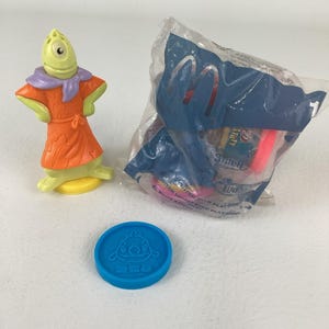 May include: A collection of children's toys featuring a green and orange figure, a blue coin with a character design, and a sealed plastic bag with various items. The bag has the McDonald's logo and text related to "Lilo & Stitch".