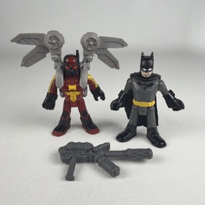 Op de afbeelding: Twee plastic actiefiguren en een grijs speelgoedwapen. Eén figuur is rood en geel met grote grijze vleugels, terwijl de andere een grijs en zwart figuur met een cape is. Het wapen is een grijs blaster-accessoire met meerdere lopen.