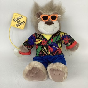 Puede incluir: Un león de peluche con gafas de sol naranjas, una camisa hawaiana con estampado floral y pantalones azules. Un letrero amarillo que dice "Bubba on Board" está adjunto al león.