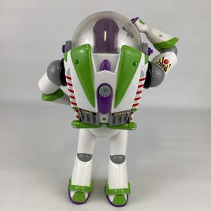 May include: A Buzz Lightyear action figure, featuring a white body with green and purple accents. The toy has a clear helmet, red and white striped wings, and the words "JET EXHAUST" and "DANGER" printed on the back.