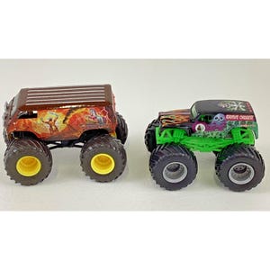 May include: Two toy monster trucks. One is brown with a graphic of a volcano and a figure. The other is black with green accents and the words "Grave Digger". Both have large tires.