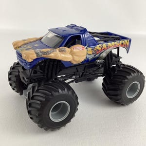 May include: A blue Hot Wheels monster truck toy with the name "SAMSON" on the side. The truck has large black tires and a design featuring muscular arms with flames. The truck is a toy vehicle.
