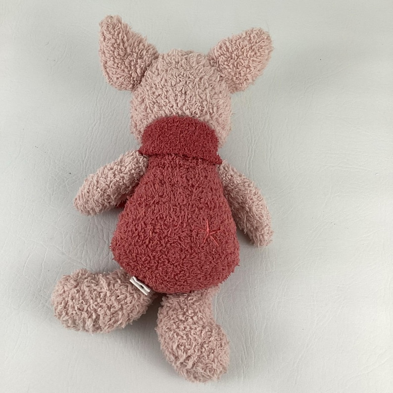 May include: A plush pig toy in shades of pink and red. The pig has large, floppy ears, a red dress, and a stitched design on the dress. The toy is made of a soft, textured material.