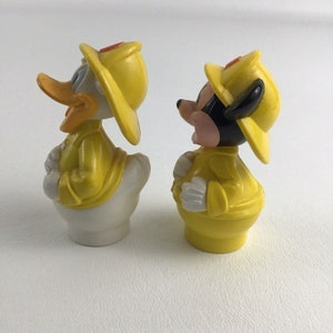May include: Two yellow plastic figurines of Mickey Mouse and Donald Duck dressed as firefighters. Both figures are wearing yellow raincoats and yellow firefighter hats with red accents. The figures are standing on a white surface.