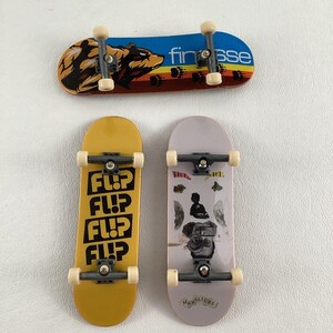 May include: Three miniature skateboards. The first has a blue deck with a bear graphic and the text "finesse". The second has a yellow deck with the text "FLIP" repeated four times. The third has a light purple deck with a graphic of a person and the text "MAX ALLURE".