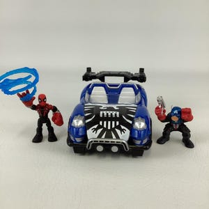 May include: A blue toy car with a black and white graphic design on the hood, flanked by two action figures. One figure is Spider-Man, and the other is Captain America holding a gun. The Spider-Man figure is holding a blue web.