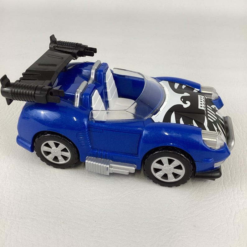 May include: A blue toy car with a black spoiler and decorative accents. The car features a white and black design on the hood and silver-colored wheels. The toy car is designed with a sporty aesthetic.