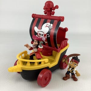 May include: A toy pirate ship with a red, black, and yellow color scheme. The ship features a skull and crossbones design on the sail and includes Captain Hook and Jake figures. The ship has red wheels and a skull figurehead.