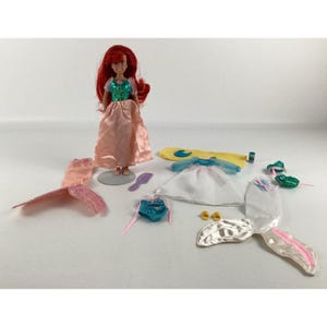 K&ouml;nnte beinhalten: Eine rothaarige Puppe, die ein rosa Kleid und ein gr&uuml;nes Oberteil tr&auml;gt. Die Puppe ist von verschiedenen Accessoires umgeben, darunter ein rosa Schwanz, ein wei&szlig;es Kleid, ein blauer Badeanzug und eine gelbe Flosse.