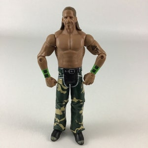 May include: A wrestling action figure with long blonde hair, a bare chest, and camouflage trousers. The figure is wearing green wristbands with the letters "DX" on them.
