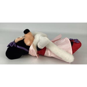 May include: A plush toy character with black ears, a peach-coloured face, and white gloved hands. The character wears a light pink satin dress with a red skirt section, a purple bow on the head, and small purple bows on the red shoes.