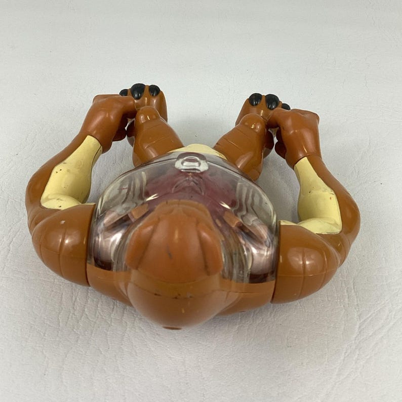 May include: A brown and cream-colored plastic action figure with a clear, rounded dome covering the chest area. The figure features articulated limbs with black-tipped hands and feet, and a smooth, molded head design with a visible circular detail.
