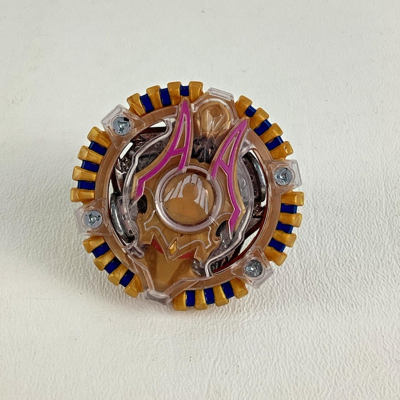 Puede incluir: Un juguete circular con un centro transparente, detalles dorados y un anillo exterior azul y dorado. El juguete presenta elementos decorativos rosas y dorados y est&aacute; asegurado con tornillos plateados. Este juguete est&aacute; dise&ntilde;ado para girar y luchar.