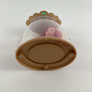 Puede incluir: Una c&uacute;pula de pl&aacute;stico transparente con una base y una parte superior en forma de corona dorada. En el interior, se ve la cabeza de una mu&ntilde;eca con pelo rojo. La base tiene los n&uacute;meros manuscritos "1050". Una peque&ntilde;a gema verde adorna la corona.