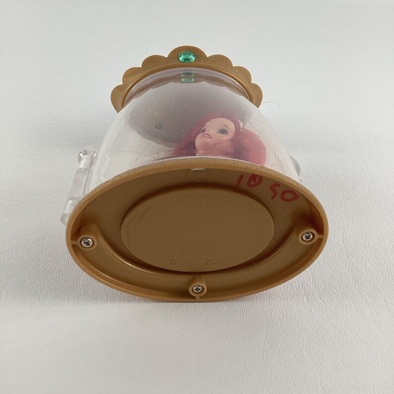 Puede incluir: Una c&uacute;pula de pl&aacute;stico transparente con una base y una parte superior en forma de corona dorada. En el interior, se ve la cabeza de una mu&ntilde;eca con pelo rojo. La base tiene los n&uacute;meros manuscritos "1050". Una peque&ntilde;a gema verde adorna la corona.
