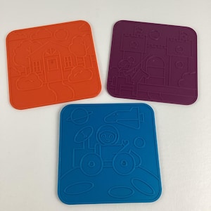 Könnte beinhalten: Drei farbige quadratische Untersetzer. Einer ist orange mit einem Hausdesign, ein anderer ist lila mit einem Burgdesign, und der letzte ist blau mit einem Weltraumdesign mit einem Astronauten und einer Rakete.