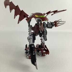 Puede incluir: Figura de acción detallada de una criatura robótica en rojo y negro. La figura tiene extremidades articuladas, alas y manos con forma de garra. La cabeza presenta una visera verde. La figura está posada sobre una superficie blanca.
