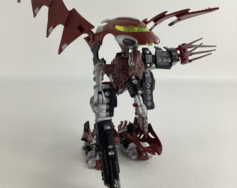 May include: A detailed action figure of a robotic creature with a red and black color scheme. The figure has articulated limbs, wings, and claw-like hands. The head features a green visor. The figure is posed on a white surface.