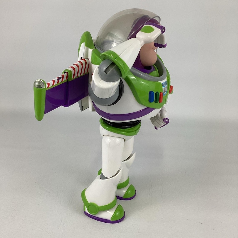 May include: A Buzz Lightyear action figure in a white, green, and purple spacesuit. The figure has a clear helmet, a green jetpack with red and white stripes, and is in a saluting pose. The toy is a collectible item.