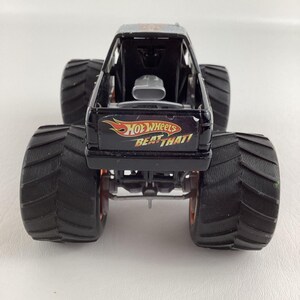 Puede incluir: Un juguete de cami&oacute;n monstruo Hot Wheels negro con neum&aacute;ticos negros grandes y detalles en naranja. El cami&oacute;n presenta el texto "HOT WHEELS BEAT THAT!" en la parte trasera. El juguete tiene un interior negro y un motor plateado.
